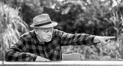 An elderly man sits with an angry face at a table in the park. He points to something with his finger. Concept unsympathetic people.