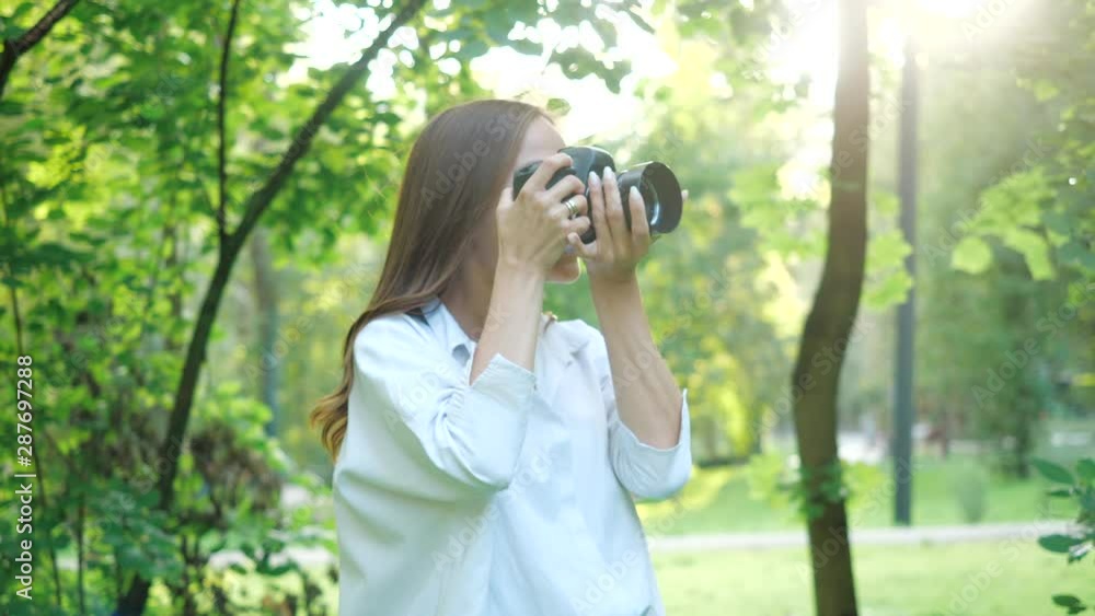 Pretty smiling girl photographer wearing white shirt is making photos with professional camera in a park on a soft background of green foliage and spraying water. Pretty smiling girl photographer wearing white shirt is making photos with professional camera in a park on a soft background of green foliage and spraying water.