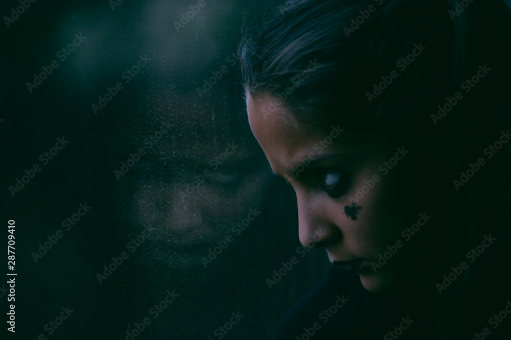 young little caucasian girl with dark makeup for Halloween holiday party looking on her reflection in window lit buy movie greenish light