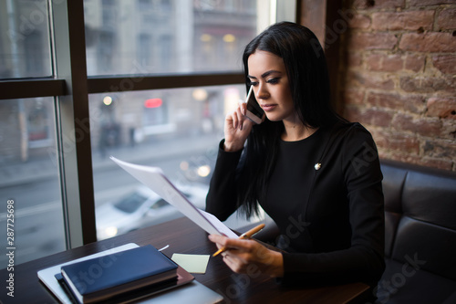 Young caucasian woman prosperous business worker holding paper document with monthly revenue report and talking with partner via cell telephone while sitting in restaurant. Manager reading resume
