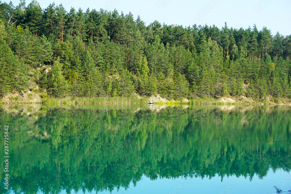 Pine forest is reflected in the surface of the green-blue water of the chalk pits in the center of Belarus, Lyuban