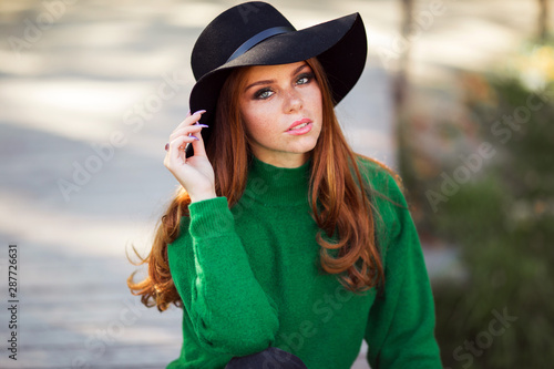 Portrait of sunshine young teen girl with red curly hair is wearing green sweater and fashion hat in autumn park.