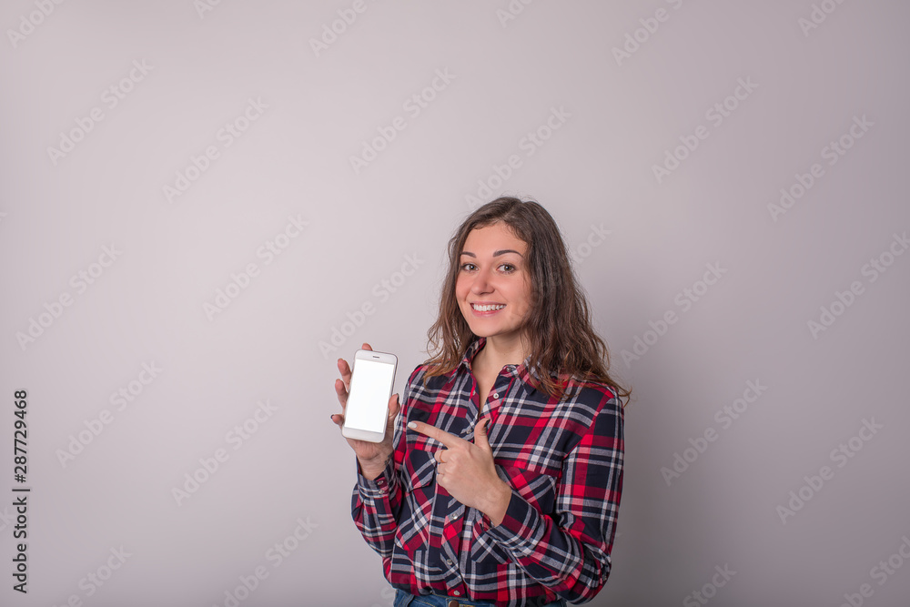 Portrait of a happy smiling attractive hipster girl in stylish casual wear shows a finger on mobile phone with empty mock up copy space screen background for promotional text message Portrait of a happy smiling attractive hipster girl in stylish casual wear shows a finger on mobile phone with empty mock up copy space screen background for promotional text message