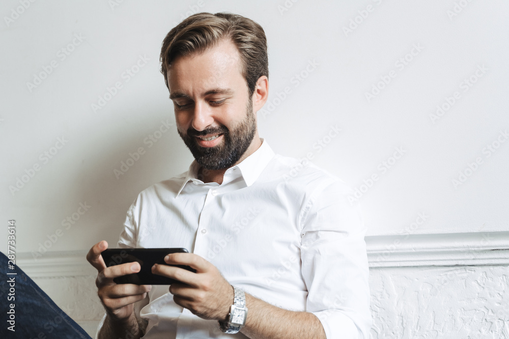 Image of happy smiling businessman playing video game on cellphone while sitting on chair in office Image of happy smiling businessman playing video game on cellphone while sitting on chair in office