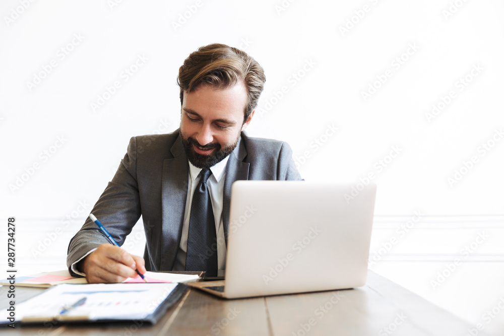 Image of happy handsome businessman working at laptop in office while writing down notes Image of happy handsome businessman working at laptop in office while writing down notes
