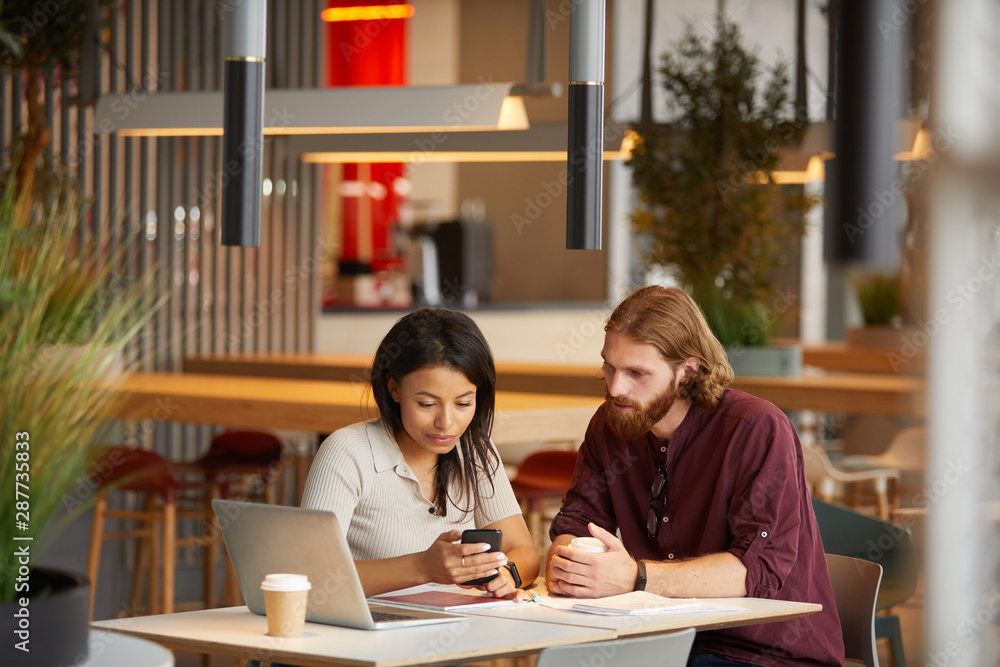 Young woman looking at mobile phone and showing something to the man while they sitting at the table with laptop and coffee drinks at meeting Young woman looking at mobile phone and showing something to the man while they sitting at the table with laptop and coffee drinks at meeting