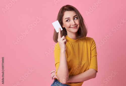 Portrait of a young woman in yellow shirt showing credit card and looking away at copy space isolated over pink background.