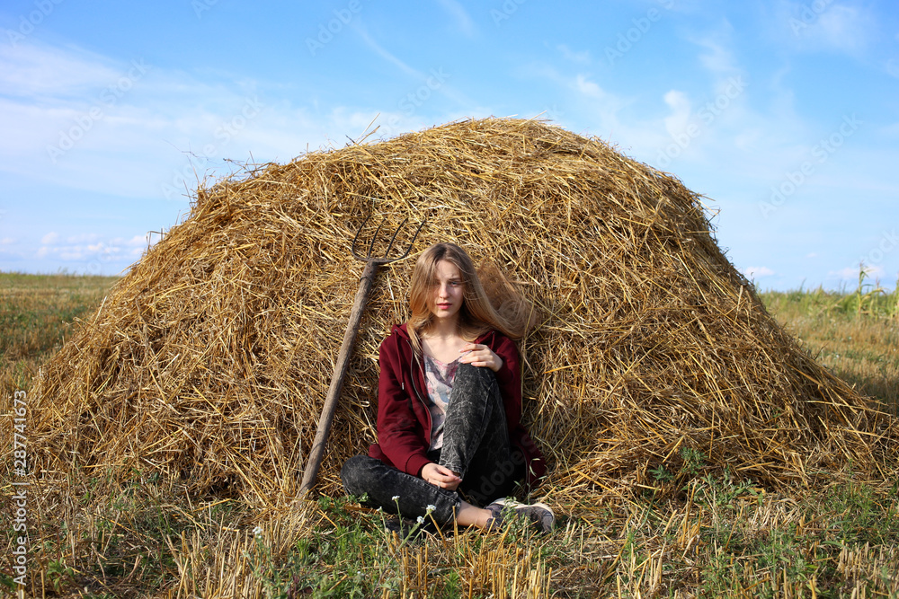 A serious, beautiful blonde girl in a burgundy sweater is sitting near a pile of dry rye hay, next to her is a pitchfork working tool. A serious, beautiful blonde girl in a burgundy sweater is sitting near a pile of dry rye hay, next to her is a pitchfork working tool.