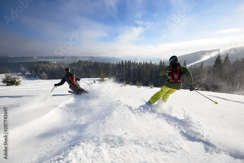 Back view of proficient male skiers in snow powder. Backcountry skiing. Using carving technique on wide open wooded hillside. Panoramic view of picturesque winter mountains and forest under blue sky. photo