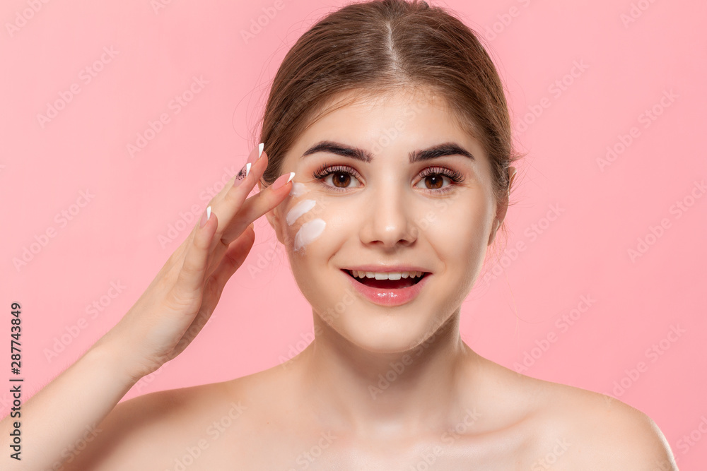 Beautiful model applyes moisturizing cosmetic product to skin holding white tube of a hyaluronic cream isolated over pink background. Concept of beauty and health treatment. Beautiful model applyes moisturizing cosmetic product to skin holding white tube of a hyaluronic cream isolated over pink background. Concept of beauty and health treatment.