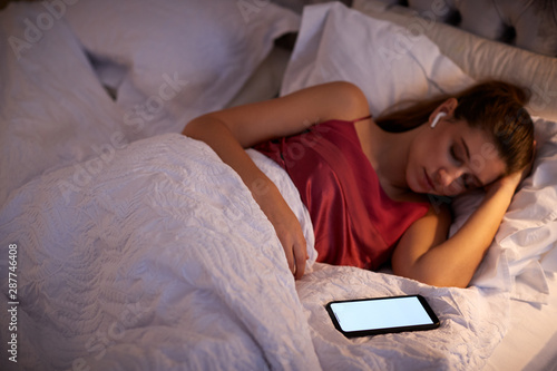 Fotografija Woman Sleeping In Bed Wearing Wireless Earphones Connected To Mobile Phone