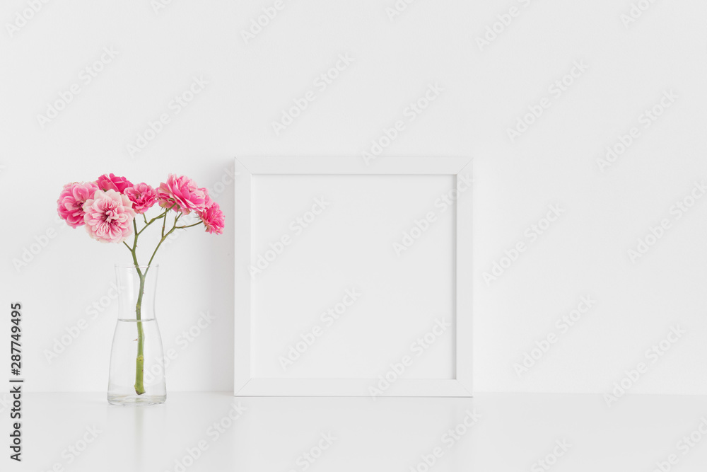 White square frame mockup with pink roses in a glass vase on a white table. White square frame mockup with pink roses in a glass vase on a white table.