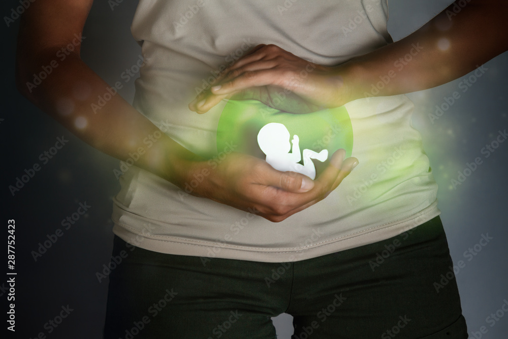 Pregnancy and abortion. The paper-cut silhouette of an embryo in a woman's hands is surrounded by a greenish glow - an allegory of the formation of a new life - obrazy, fototapety, plakaty 