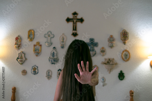 Young girl with long mane in front of her face sitting on the bed shows the palm of her hand in a dark room, in the background there is a cross and several religious elements photo