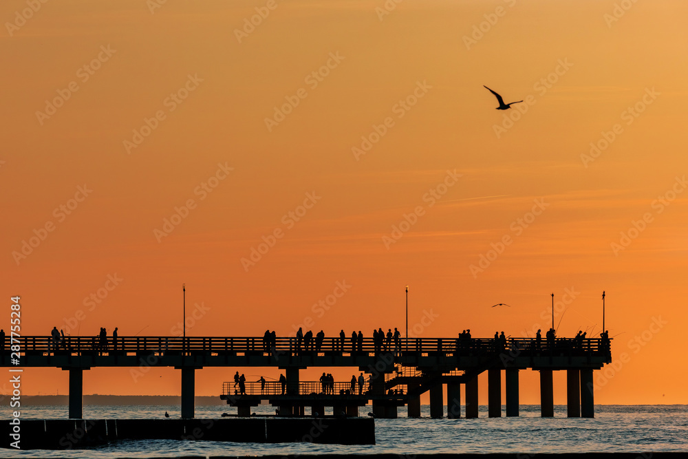 silhouettes of people walking on a pier by the sea against a sunset orange sky