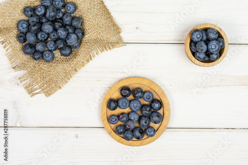 Lot of whole fresh blue bilberry in tiny wooden bowl on round bamboo coaster on natural sackcloth flatlay on white wood