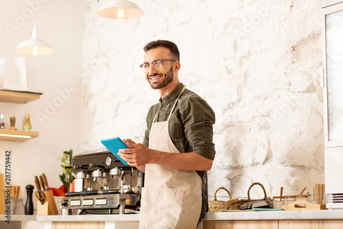 Cheerful owner of coffee shop smiling while holding little tablet