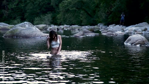 An attractive young brunette woman swimming in a pristine forest river with a creepy voyeur photographing her from afar. photo