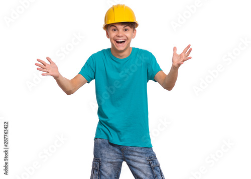 Portrait of smiling teen boy wearing yellow hard hat, isolated on white background. Beautiful student caucasian young teenager in helmet looking at camera. Cheerful child with raised hands shouting.