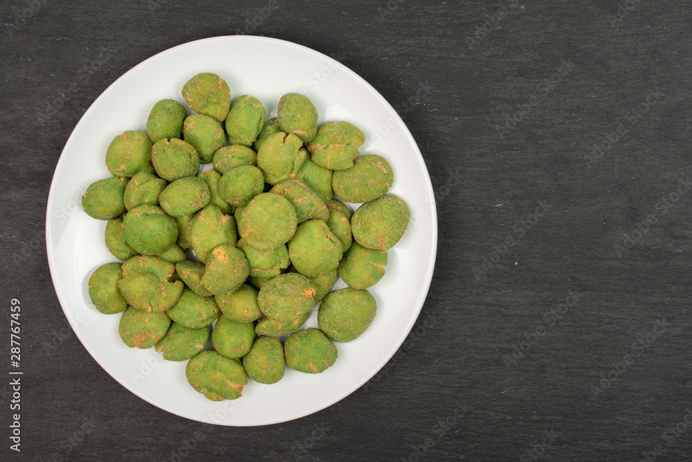 Lot of whole spicy green wasabi peanut on white ceramic plate flatlay on grey stone Lot of whole spicy green wasabi peanut on white ceramic plate flatlay on grey stone
