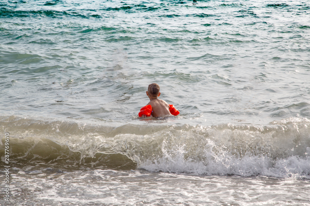 boy in red inflatable sleeves walks along the wave