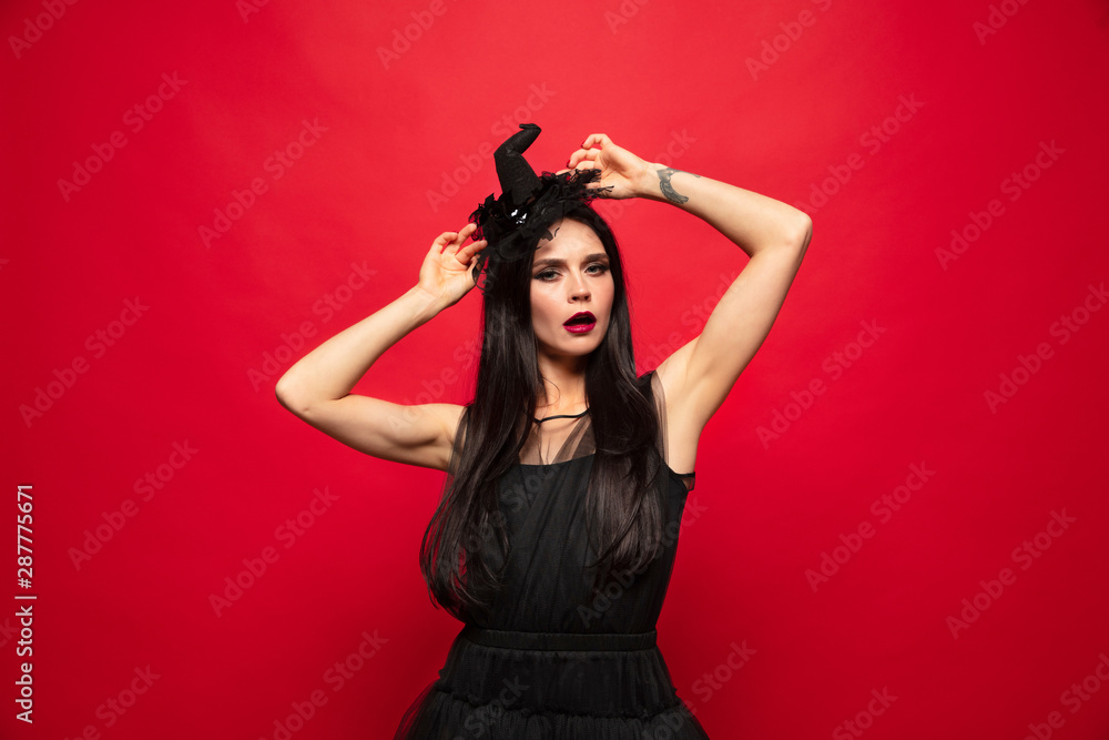 Young brunette woman in black hat and costume on red background. Attractive caucasian female model. Halloween, black friday, cyber monday, sales, autumn concept. Copyspace. Dancing, posing charming. Young brunette woman in black hat and costume on red background. Attractive caucasian female model. Halloween, black friday, cyber monday, sales, autumn concept. Copyspace. Dancing, posing charming.