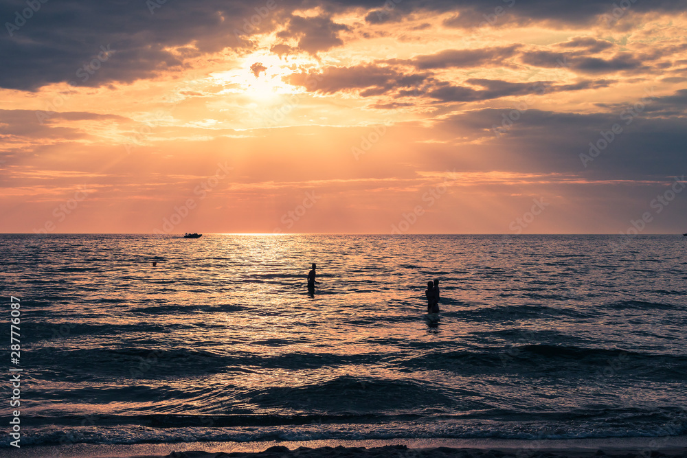 Beautiful landscape with three silhouettes swimming in the sea during sunset under the cloudy sky.