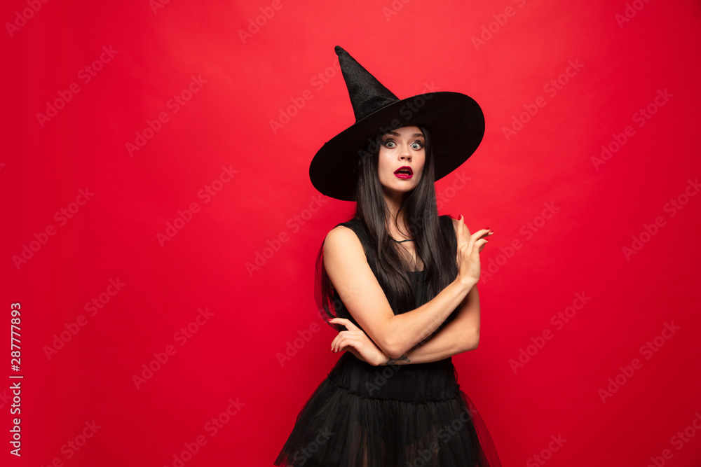 Young brunette woman in black hat and costume on red background. Attractive caucasian female model. Halloween, black friday, cyber monday, sales, autumn concept. Copyspace. Pointing up. Young brunette woman in black hat and costume on red background. Attractive caucasian female model. Halloween, black friday, cyber monday, sales, autumn concept. Copyspace. Pointing up.