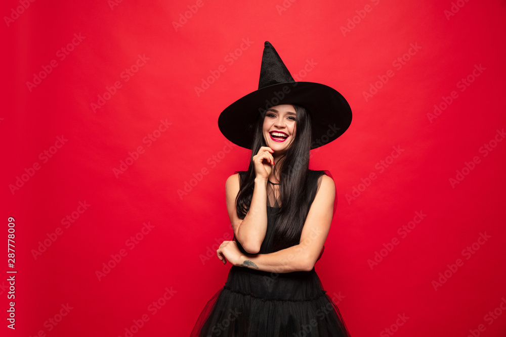 Young brunette woman in black hat and costume on red background. Attractive caucasian female model. Halloween, black friday, cyber monday, sales, autumn concept. Happy smiling. Young brunette woman in black hat and costume on red background. Attractive caucasian female model. Halloween, black friday, cyber monday, sales, autumn concept. Happy smiling.