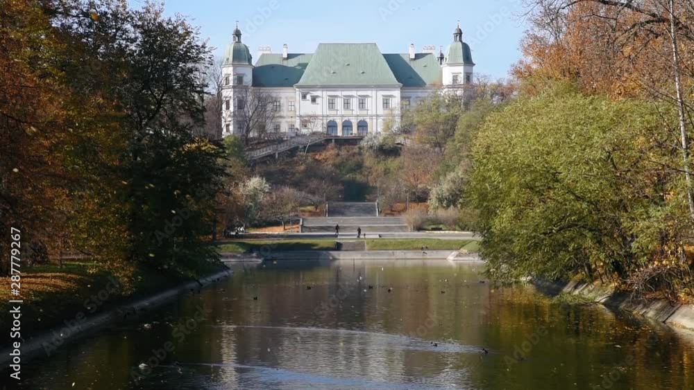 City park Royal Lazienki with pond or lake and Ujazdowski Castle on background with blue sky, trees and fallen leaves at autumn. Warsaw, Poland, Europe. Camera move from bottom to top City park Royal Lazienki with pond or lake and Ujazdowski Castle on background with blue sky, trees and fallen leaves at autumn. Warsaw, Poland, Europe. Camera move from bottom to top