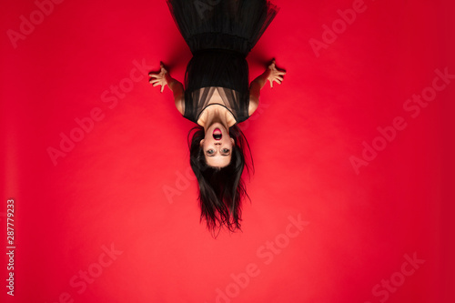 Young brunette woman in black hat and costume on red background. Attractive caucasian female model. Halloween, black friday, cyber monday, sales, autumn concept. Scary screaming upside down.