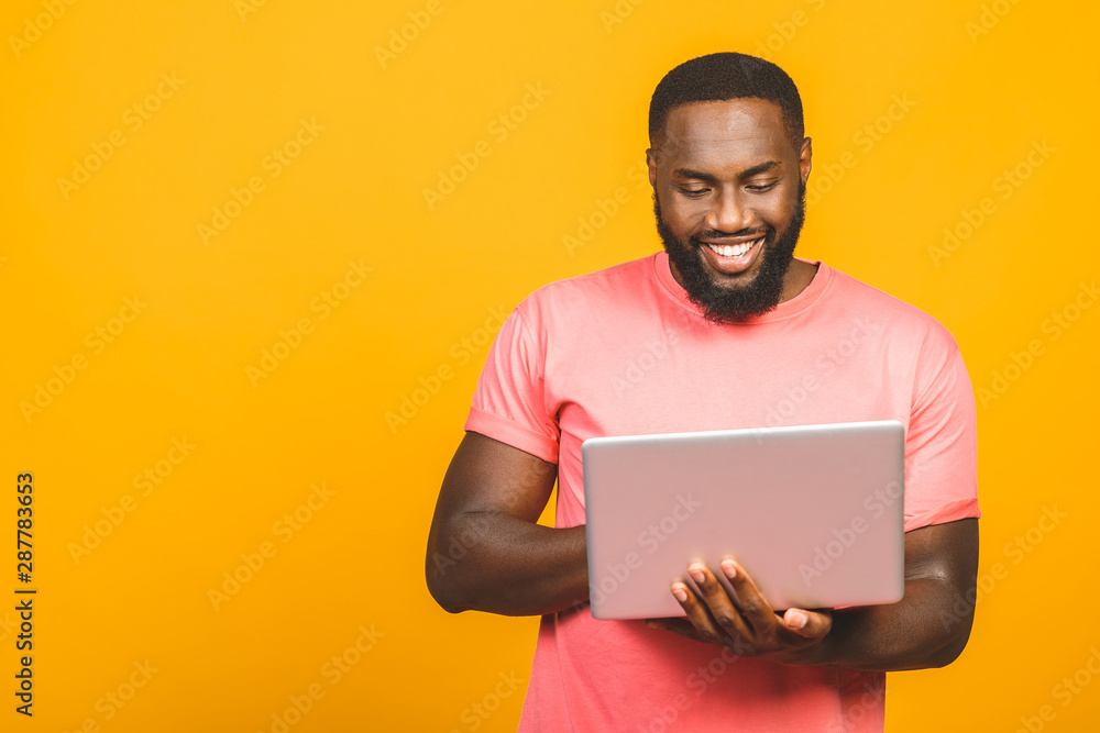 Young smiling african american man standing and using laptop computer isolated over yellow background.