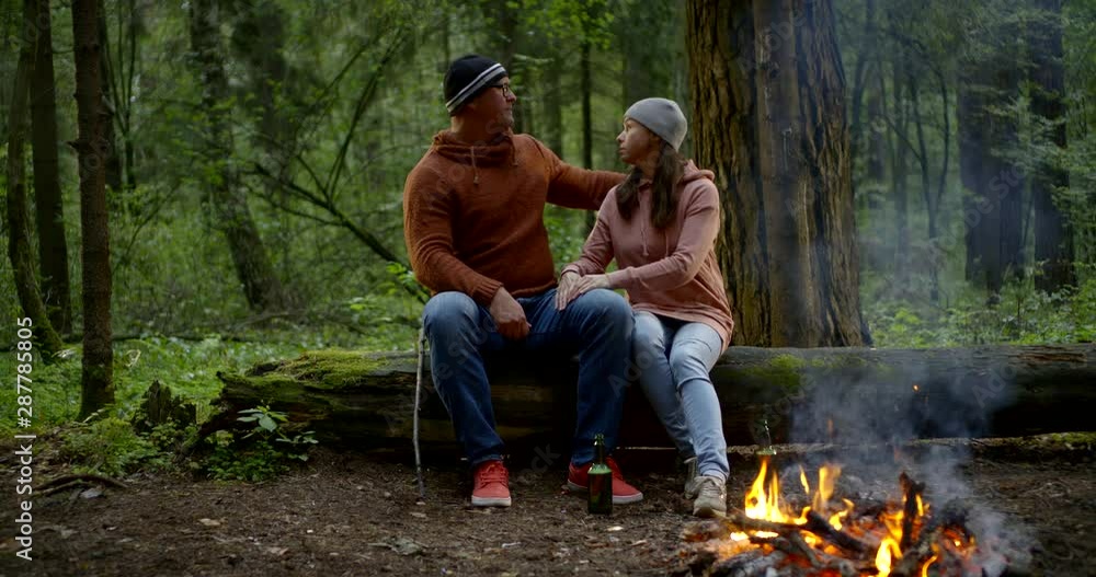 A middle-aged couple are in the forest and they are sitting near a burning fire on a log, laughing, talking, hugging, kissing. They have hats on their heads. A middle-aged couple are in the forest and they are sitting near a burning fire on a log, laughing, talking, hugging, kissing. They have hats on their heads.