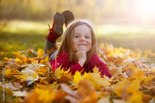Little funny girl lies on yellow leaves in the forest. Child on a walk in the autumn park. Preschool girl on the street