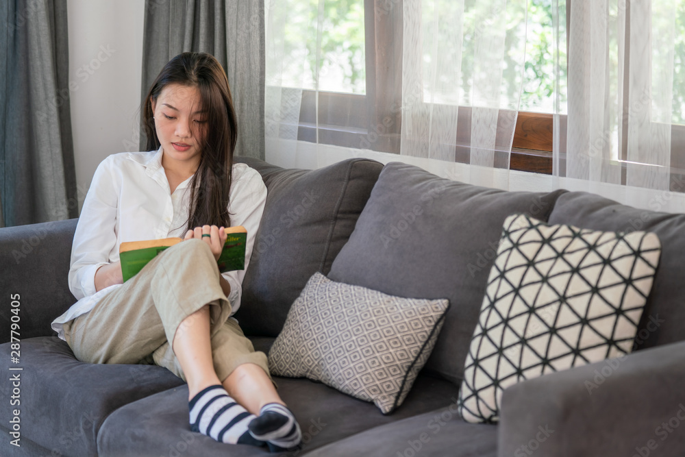 Selective focus of attractive young asian woman lying down on sofa in living room and reading a book alone. Beautiful teenage girl resting and enjoying to read magazine at home in weekend. Selective focus of attractive young asian woman lying down on sofa in living room and reading a book alone. Beautiful teenage girl resting and enjoying to read magazine at home in weekend.