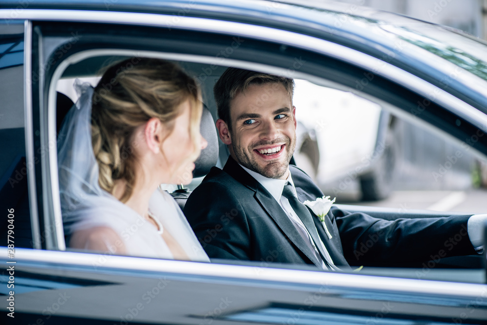 selective focus of attractive bride in bridal veil and bridegroom smiling in car