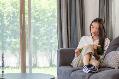 Selective focus of attractive young asian woman lying down on sofa in living room and reading a book alone. Beautiful teenage girl resting and enjoying to read magazine at home in weekend.