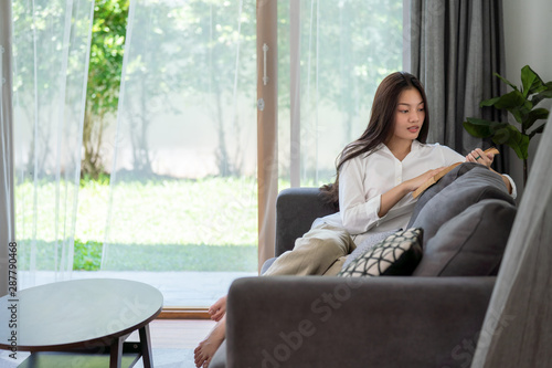 Selective focus of attractive young asian woman lying down on sofa in living room and reading a book alone. Beautiful teenage girl resting and enjoying to read magazine at home in weekend.