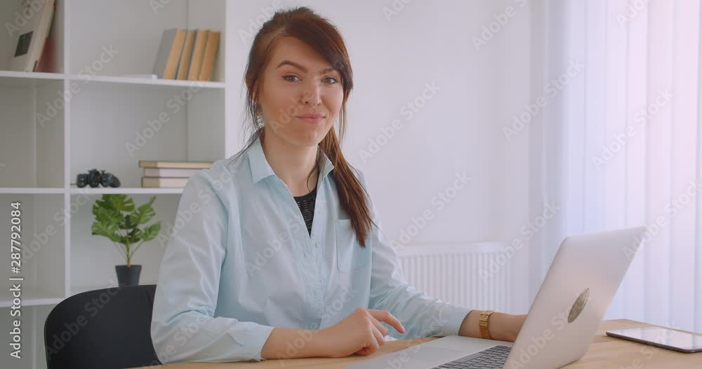 Closeup portrait of young attractive caucasian female doctor using laptop looking at camera smiling cheerfully indoors in office Closeup portrait of young attractive caucasian female doctor using laptop looking at camera smiling cheerfully indoors in office