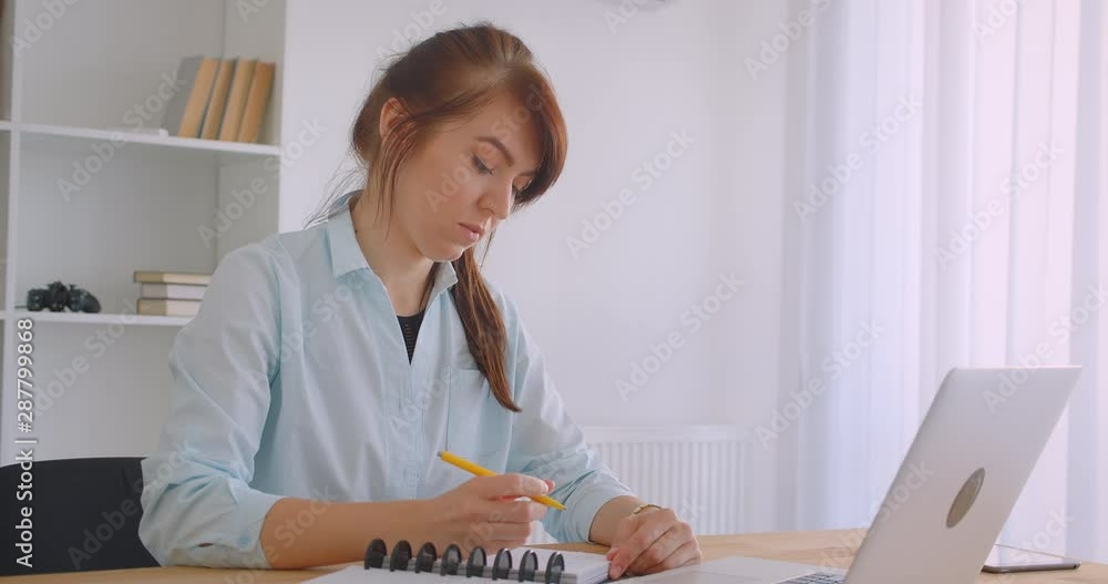 Closeup portrait of young caucasian female doctor in coat using laptop taking notes indoors in office Closeup portrait of young caucasian female doctor in coat using laptop taking notes indoors in office