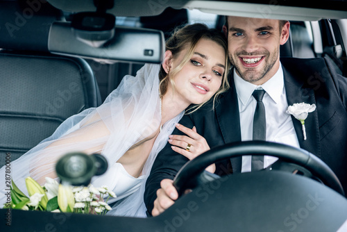 attractive bride in bridal veil hugging handsome bridegroom in car