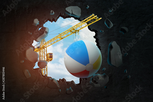 3d rendering of hoisting crane carrying beach ball and breaking hole in black wall with blue sky seen through. photo