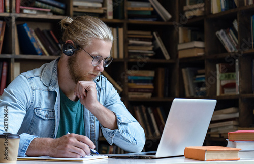 Focused serious young man wear wireless headset look at laptop screen make notes study e learning in library watching webinar training online course video calling, distance education software concept photo