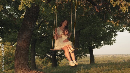 mother and baby ride on rope swing on an oak branch in forest. Mom shakes her daughter on swing under a tree in sun. Girl laughs, rejoices. Family fun in park, in nature. warm summer day.