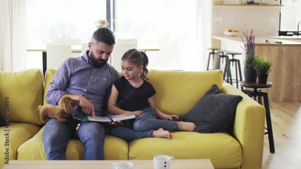 Mature father with small daughter sitting on sofa indoors, reading a book. Mature father with small daughter sitting on sofa indoors, reading a book.