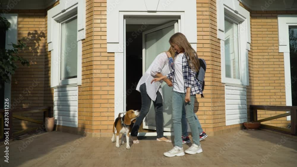 Small cute brother and sister with backpacks leaving the house and going to school. Smiling mother waving hand. Dog meets children and wagging its tail. Happy loving family concept Small cute brother and sister with backpacks leaving the house and going to school. Smiling mother waving hand. Dog meets children and wagging its tail. Happy loving family concept