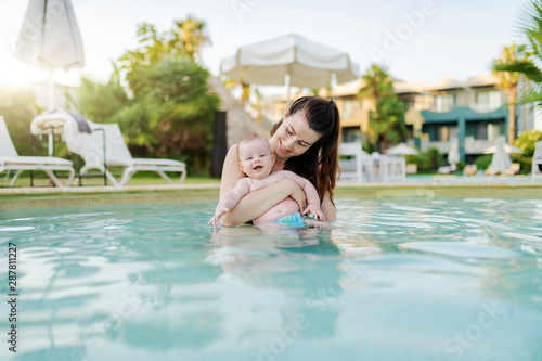 Beautiful Caucasian mother standing in swimming pool and holding her 6 months old son. Baby enjoying and smiling.