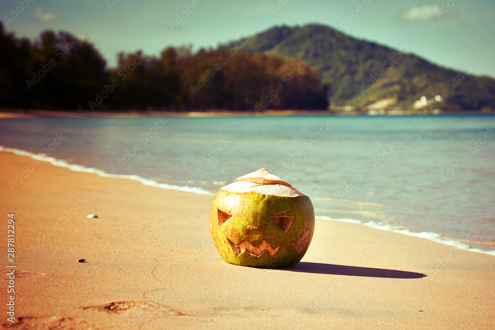 Obraz premium The symbol of Halloween. Instead of a pumpkin, a coconut with a face carved on it like a jack lantern. Fresh green coconut lies on the beach against the backdrop of a tropical island. Warm toned