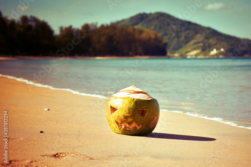 The symbol of Halloween. Instead of a pumpkin, a coconut with a face carved on it like a jack lantern. Fresh green coconut lies on the beach against the backdrop of a tropical island. Warm toned