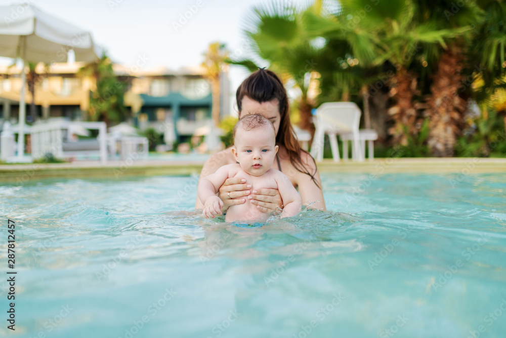 Curious Caucasian 6 months old baby boy learning how to swim at swimming pool. Mother holding her son. First time at pool concept.